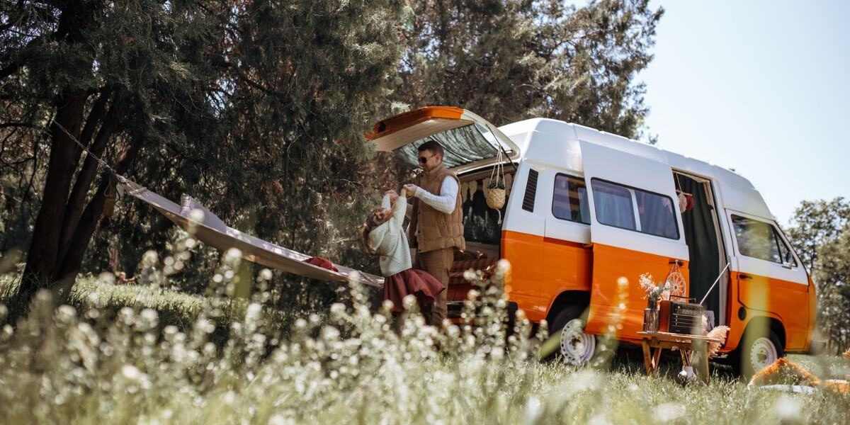 Oranssi retkeilyauto niityllä, isä ja tytär leikkimässä sen takaluukulla